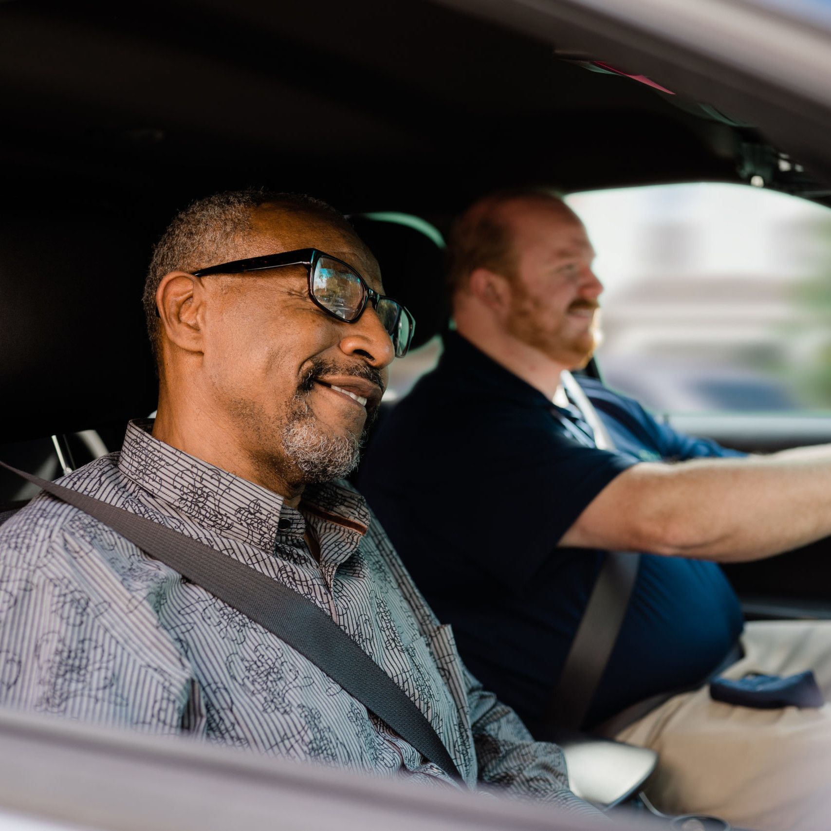 Black man with glasses is seated in the passenger seat as white man with a short beard drives.