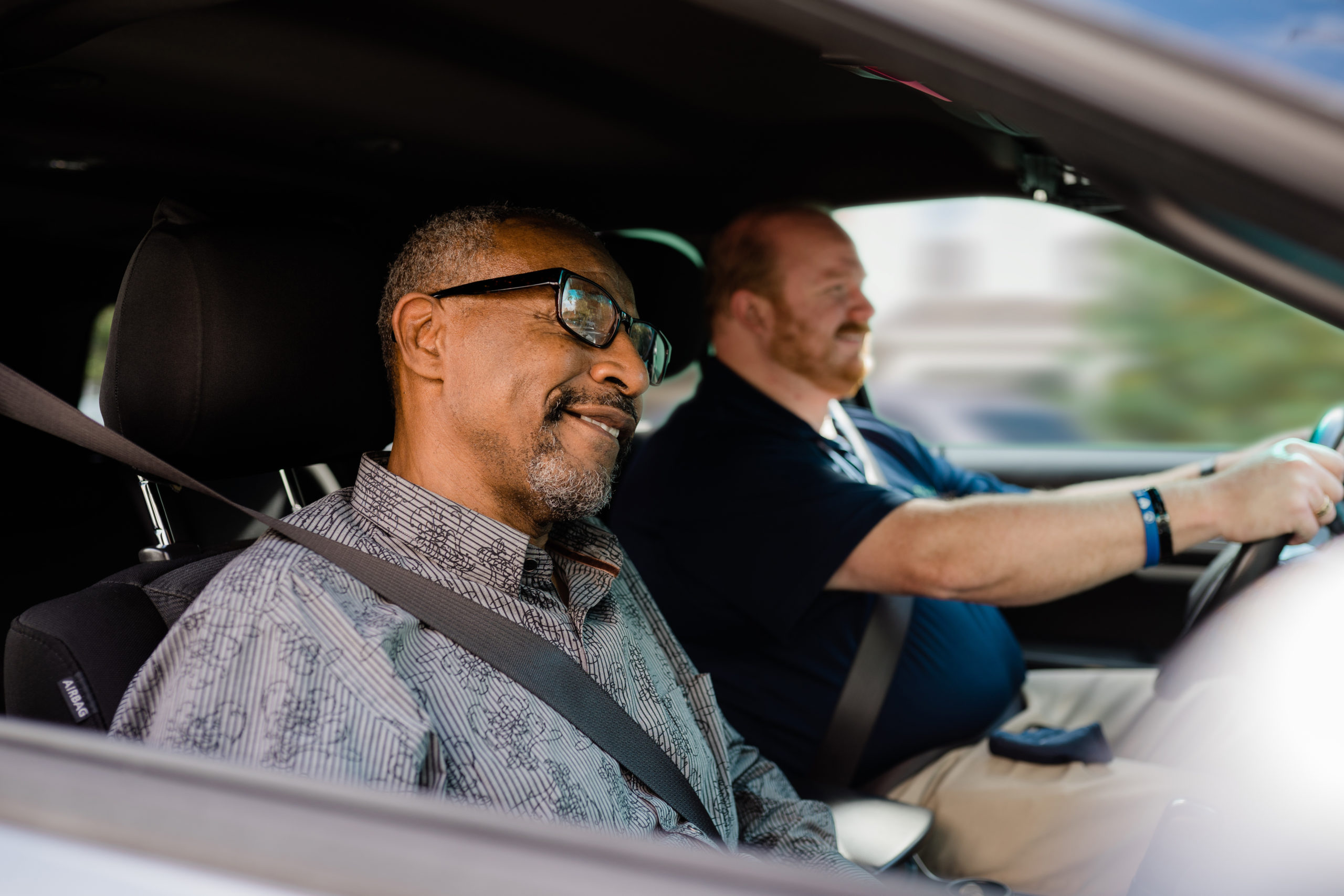 Black man with glasses is seated in the passenger seat as white man with a short beard drives.