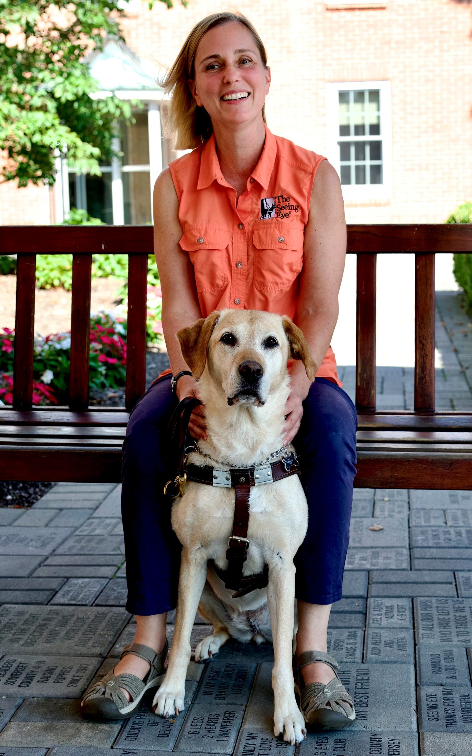 Melissa Allman sits on a bench, wind in her hair, with a smile on her face. She is wearing a slamon tank top and blue pants. Her service dog, Luna, sits at attention between her knees. Luna is a Golden Retriever, wearing a brown harness