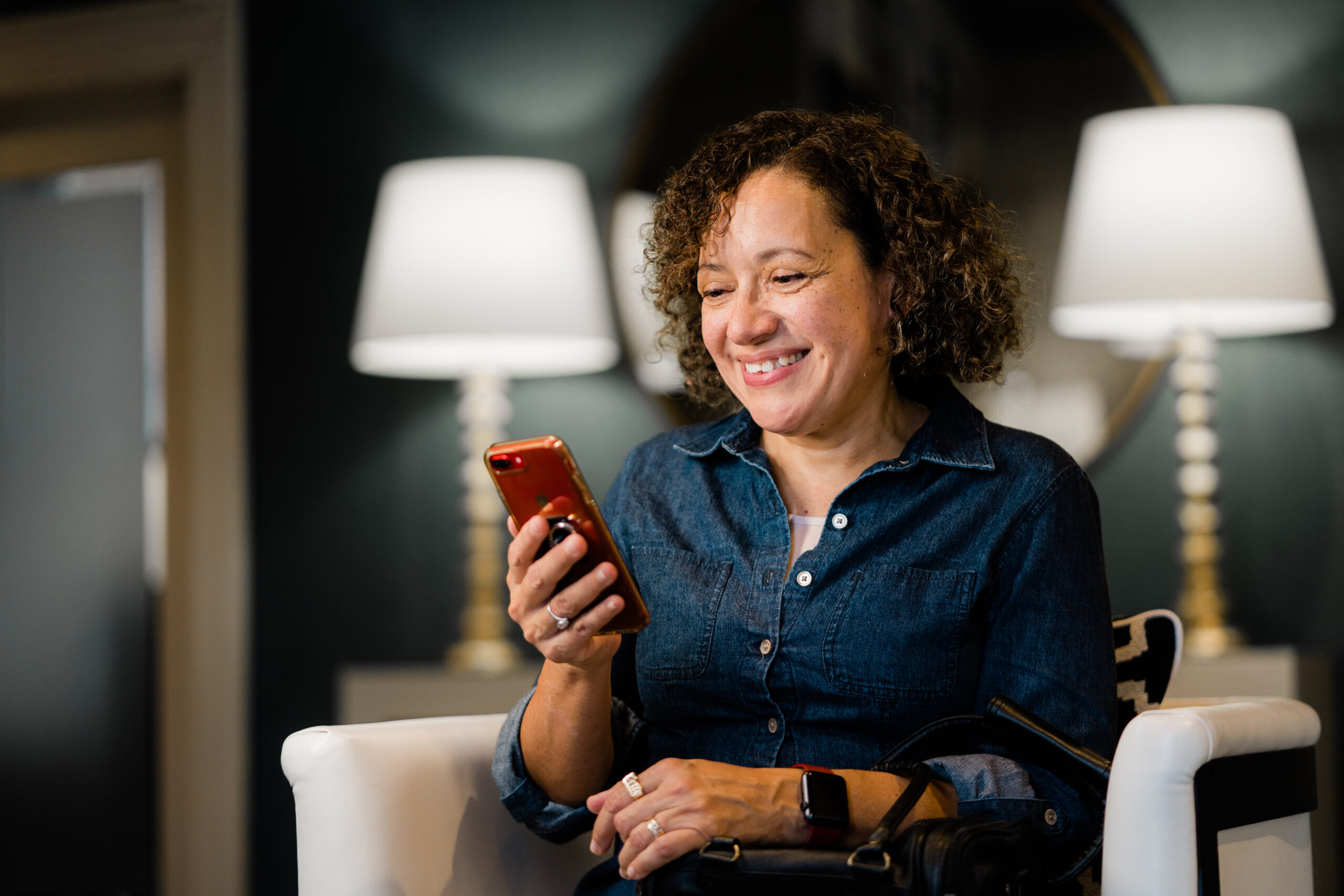 A woman is sitting down while using her phone. She is smiling.